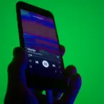 A hand holds a smartphone displaying a music player app with a song titled "Change" by "Like Calling Waves" against a green background.