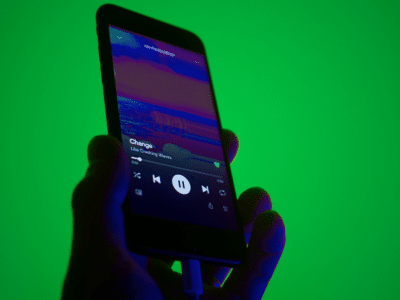 A hand holds a smartphone displaying a music player app with a song titled "Change" by "Like Calling Waves" against a green background.