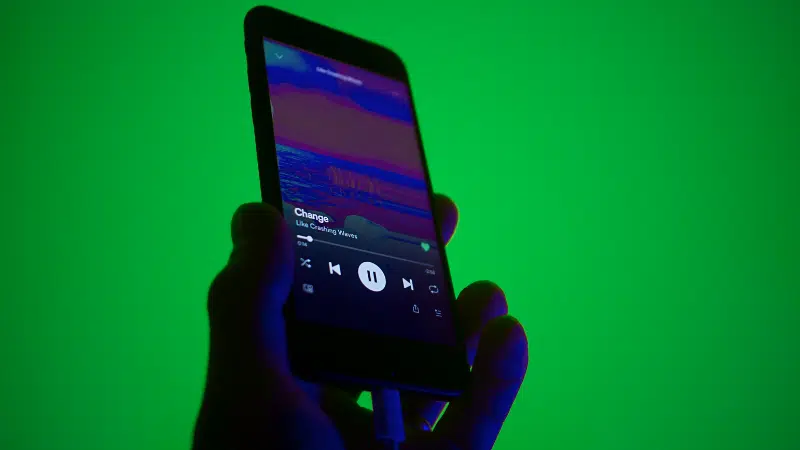 A hand holds a smartphone displaying a music player app with a song titled "Change" by "Like Calling Waves" against a green background.