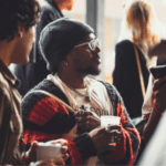 A group of people socializing indoors, with focus on a person wearing glasses, a black beanie, and a striped sweater, holding a beverage.