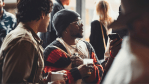 A group of people socializing indoors, with focus on a person wearing glasses, a black beanie, and a striped sweater, holding a beverage.