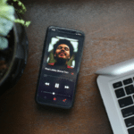 A smartphone displaying the Apple Music app lies next to a partially visible laptop and a potted plant on a wooden surface.