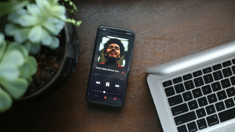 A smartphone displaying the Apple Music app lies next to a partially visible laptop and a potted plant on a wooden surface.