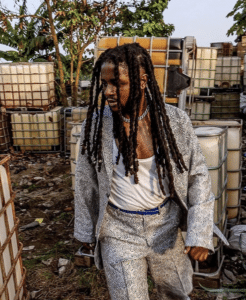 A person with long dreadlocks, wearing a patterned suit and a white tank top, walks through an area with stacked containers and lush vegetation in the background, capturing the essence of unfiltered Africa.