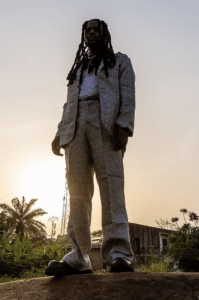 A person with long hair stands outdoors wearing a patterned suit with a white shirt, under the golden hues of a setting sun and partly cloudy sky, capturing the essence of unfiltered Africa.