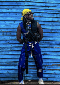 Person wearing blue pants, a sleeveless navy top, and a yellow bandana stands unfiltered against a blue wooden wall. They have a cross-body bag and sunglasses.