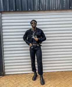 Person standing in front of a corrugated metal garage door, wearing an unfiltered black jacket, black pants, black hat, dark sunglasses, and sleek black boots.