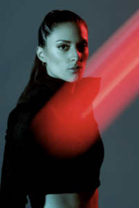 A woman with long hair pulled back is shown against a dark background with red light streaks crossing in front of her. She is wearing a high-collared, dark outfit.