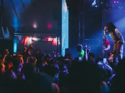 A crowded concert with a performer on stage, illuminated by colorful lights, as the audience energetically responds.