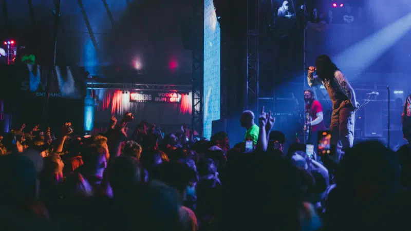 A crowded concert with a performer on stage, illuminated by colorful lights, as the audience energetically responds.