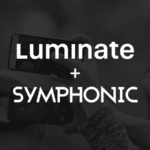 A dark background with the text "Luminate + Symphonic" in bold white letters. Multiple hands holding smartphones, each screen glowing softly, are visible in the background.