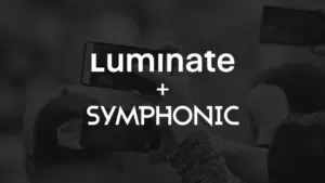 A dark background with the text "Luminate + Symphonic" in bold white letters. Multiple hands holding smartphones, each screen glowing softly, are visible in the background.