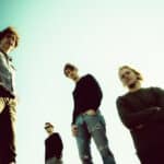 Four individuals, heralding new music, stand outdoors against a clear sky, photographed from a low angle.