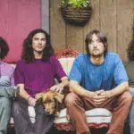 Four men with long hair and casual clothing sit on an ornate couch, embodying the spirit of rock anthems. A brown dog sits comfortably between two of the men, while a potted plant hangs behind them.