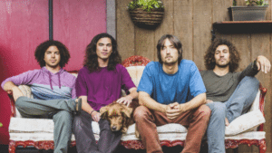Four men with long hair and casual clothing sit on an ornate couch, embodying the spirit of rock anthems. A brown dog sits comfortably between two of the men, while a potted plant hangs behind them.