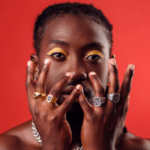 A person poses unfiltered in front of a red background with hands raised to their face, showcasing multiple rings and eye makeup.