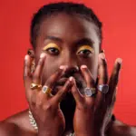 A person poses unfiltered in front of a red background with hands raised to their face, showcasing multiple rings and eye makeup.