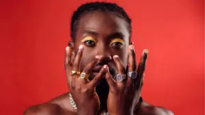 A person poses unfiltered in front of a red background with hands raised to their face, showcasing multiple rings and eye makeup.