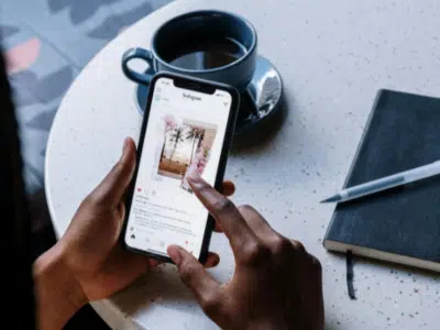 A person uses a smartphone to view an Instagram post of palm trees while sitting at a table with a black notebook, pen, and cup of coffee, effortlessly navigating their social media feed.