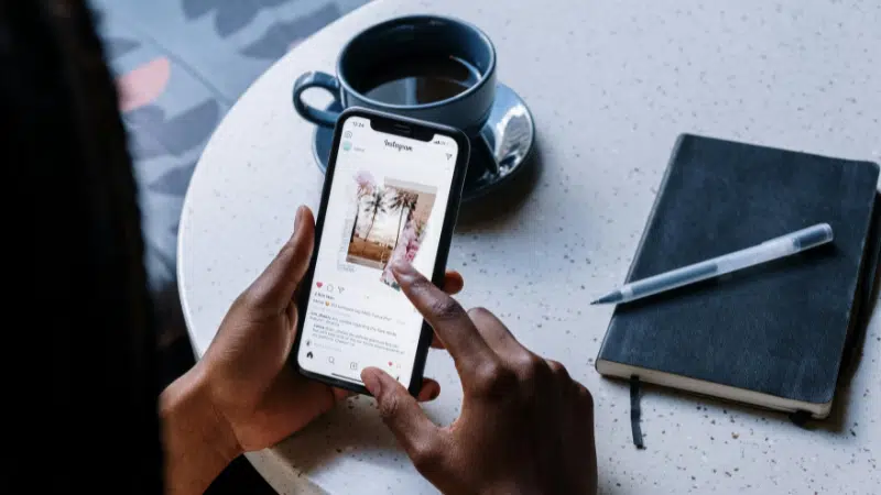 A person uses a smartphone to view an Instagram post of palm trees while sitting at a table with a black notebook, pen, and cup of coffee, effortlessly navigating their social media feed.