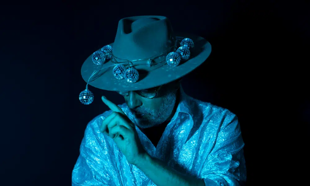 Person wearing a hat decorated with small disco balls, dressed in a shiny textured shirt, with a finger raised near their face, all under blue lighting that hints at new music vibes.