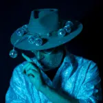 Person wearing a hat decorated with small disco balls, dressed in a shiny textured shirt, with a finger raised near their face, all under blue lighting that hints at new music vibes.