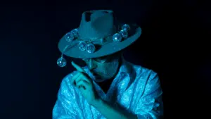 Person wearing a hat decorated with small disco balls, dressed in a shiny textured shirt, with a finger raised near their face, all under blue lighting that hints at new music vibes.