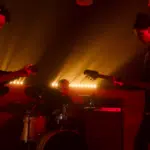 Three musicians perform on stage with dramatic backlighting; one plays guitar, another plays bass, and the third is on drums. The scene has a red hue, creating an intense atmosphere reminiscent of new music you'll discover on SubmitHub.