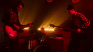 Three musicians perform on stage with dramatic backlighting; one plays guitar, another plays bass, and the third is on drums. The scene has a red hue, creating an intense atmosphere reminiscent of new music you'll discover on SubmitHub.