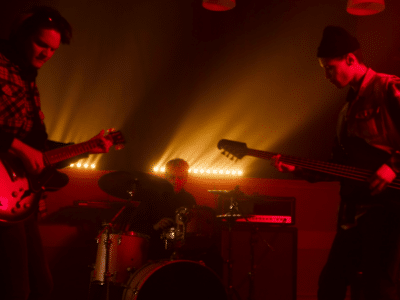 Three musicians perform on stage with dramatic backlighting; one plays guitar, another plays bass, and the third is on drums. The scene has a red hue, creating an intense atmosphere reminiscent of new music you'll discover on SubmitHub.