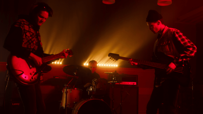 Three musicians perform on stage with dramatic backlighting; one plays guitar, another plays bass, and the third is on drums. The scene has a red hue, creating an intense atmosphere reminiscent of new music you'll discover on SubmitHub.