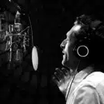 A person wearing headphones is speaking into a large microphone in a dimly lit setting, possibly recording new music.