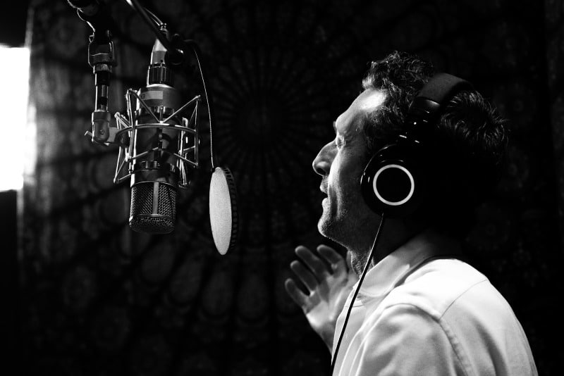 A person wearing headphones is speaking into a large microphone in a dimly lit setting, possibly recording new music.