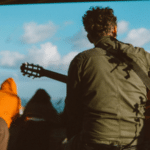 A person with curly hair, wearing a green jacket, is seen from behind playing an acoustic guitar outdoors with people in the background—perfectly capturing a moment worthy of social media.