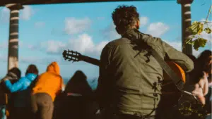 A person with curly hair, wearing a green jacket, is seen from behind playing an acoustic guitar outdoors with people in the background—perfectly capturing a moment worthy of social media.