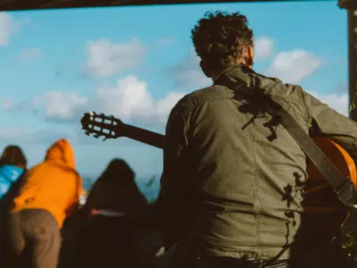 A person with curly hair, wearing a green jacket, is seen from behind playing an acoustic guitar outdoors with people in the background—perfectly capturing a moment worthy of social media.