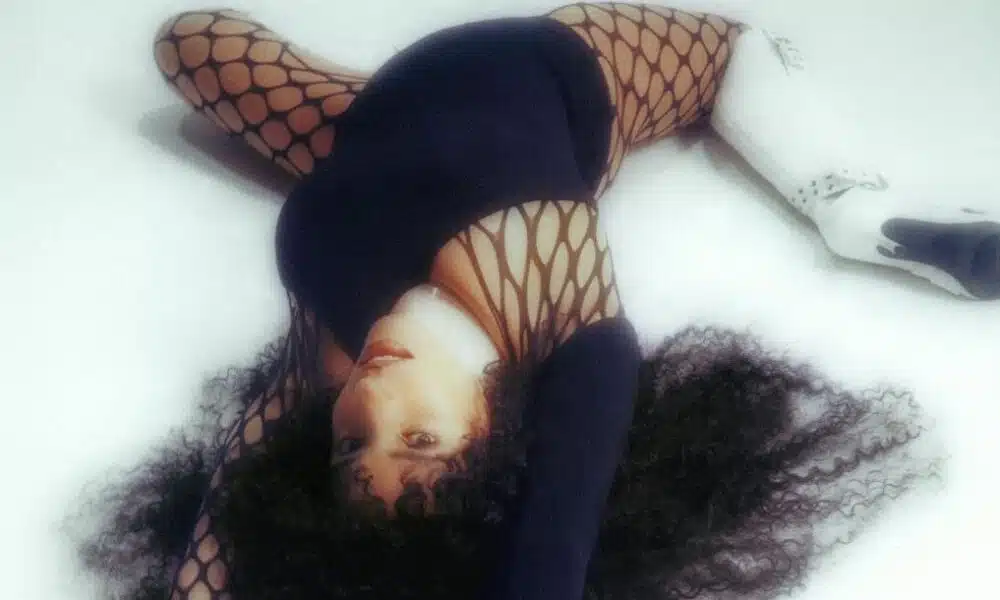 Person lying on the floor, wearing a black outfit with fishnet pattern and white, high-heeled boots. Curly hair spread on the ground and arms extended, as if silently vibing to new music.