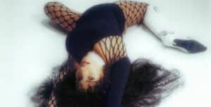 Person lying on the floor, wearing a black outfit with fishnet pattern and white, high-heeled boots. Curly hair spread on the ground and arms extended, as if silently vibing to new music.