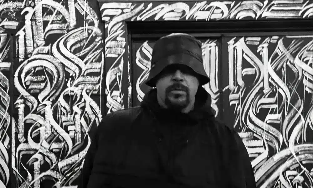 A person wearing a bucket hat and dark clothing stands before a wall adorned with abstract graffiti art, seemingly inspired by the energy of new music.