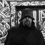 A person wearing a bucket hat and dark clothing stands before a wall adorned with abstract graffiti art, seemingly inspired by the energy of new music.