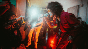 A performer in red sings passionately into a microphone, while photographers capture the moment amidst vivid stage lighting in a crowded room, creating an unforgettable scene ripe for advertising.