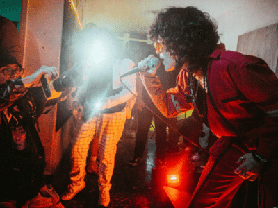 A performer in red sings passionately into a microphone, while photographers capture the moment amidst vivid stage lighting in a crowded room, creating an unforgettable scene ripe for advertising.