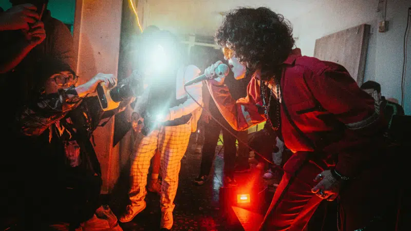 A performer in red sings passionately into a microphone, while photographers capture the moment amidst vivid stage lighting in a crowded room, creating an unforgettable scene ripe for advertising.