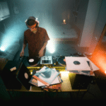 A DJ with headphones stands at a turntable setup in a dimly lit room, surrounded by vinyl records and speakers. Two spotlights illuminate the space from opposite corners.