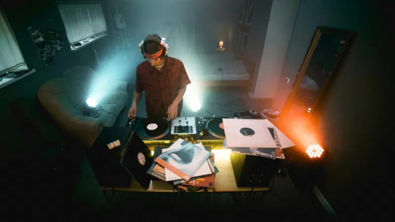 A DJ with headphones stands at a turntable setup in a dimly lit room, surrounded by vinyl records and speakers. Two spotlights illuminate the space from opposite corners.