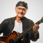 An elderly man wearing a black leather jacket and cap plays an acoustic guitar, smiling as if reminiscing about his latest songwriting contest victory against a plain white background.