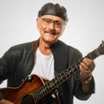 An elderly man wearing a black leather jacket and cap plays an acoustic guitar, smiling as if reminiscing about his latest songwriting contest victory against a plain white background.