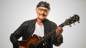 An elderly man wearing a black leather jacket and cap plays an acoustic guitar, smiling as if reminiscing about his latest songwriting contest victory against a plain white background.