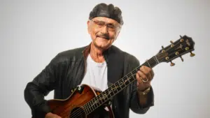 An elderly man wearing a black leather jacket and cap plays an acoustic guitar, smiling as if reminiscing about his latest songwriting contest victory against a plain white background.