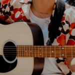 A person wearing a patterned shirt strums an acoustic guitar outdoors, as if playing for nature's royalties.